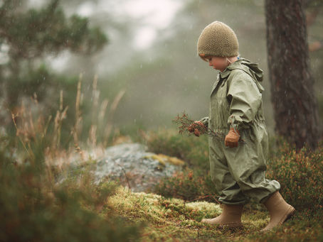 A young child wearing full sage green Nordic rain gear — including a waterproof playsuit, matching rain hat, gloves, and rubber boots — explores a misty forest.