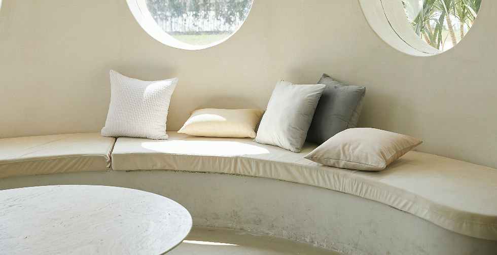 Un sofá curvo beige con cojines blancos y grises en una sala iluminada por ventanas redondas. Ambiente luminoso y relajante.