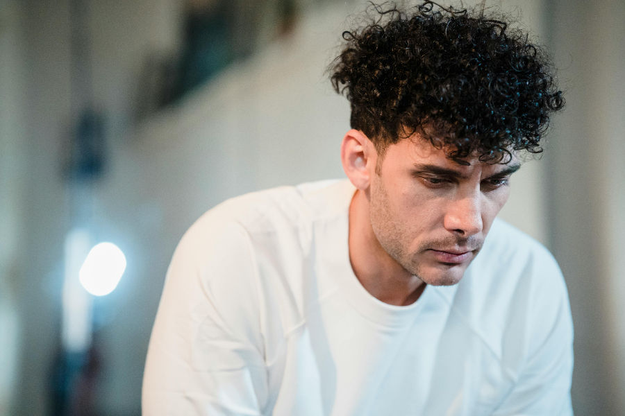 Hombre con cabello rizado mira pensativo hacia abajo, lleva camiseta blanca. Fondo desenfocado con luz brillante. Ambiente introspectivo.