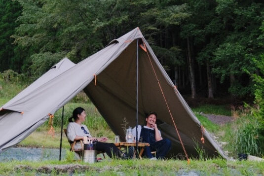森の中の草地で、グレーのタープテントの下に座る2人がキャンプを楽しみ、笑顔でくつろいでいる。