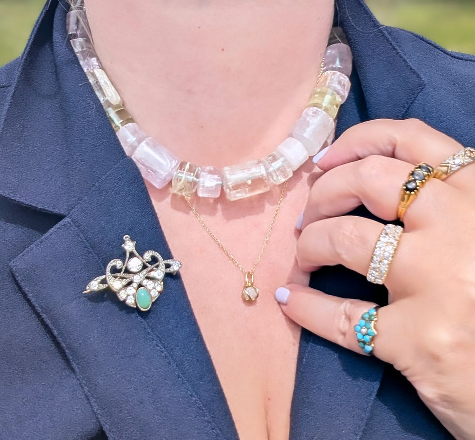 A person in a blue blazer adorned with diamond brooch. Wearing a clear crystal necklace, gold pendant, and rings with turquoise and black gems.