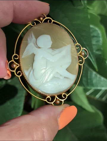 Thumbnail: a ornate yellow gold antique brooch set with a carved shell cameo of cupid writing love letters
