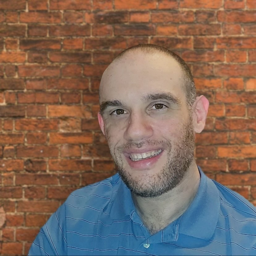Etai, a German TV Club host, wearing a blue shirt, sitting in front of a brick wall