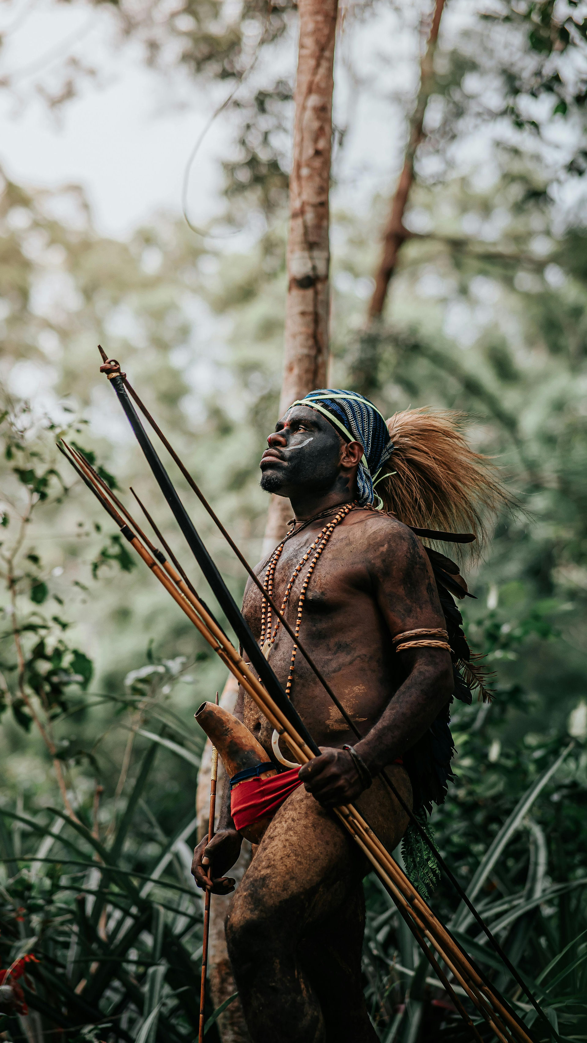 a tribal male with bow and arrow in the jungle wearing a head scarf and bushy hair and beads and face paint 