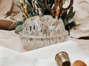 Um suporte de vidro transparente e facetado abriga várias tesouras vintage com cabos decorativos em tons de bronze e prata. Em primeiro plano, sobre um tecido branco, estão um dedal e um carretel de linha preta. O fundo é composto por papel pardo amassado e folhas verdes, misturando elementos rústicos e delicados.