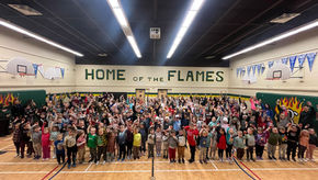 Students of various ages enthusiastically take part in the Great Big Crunch inside a school gymnasium. They bite into apples at the same time, surrounded by colourful posters and supportive educators.