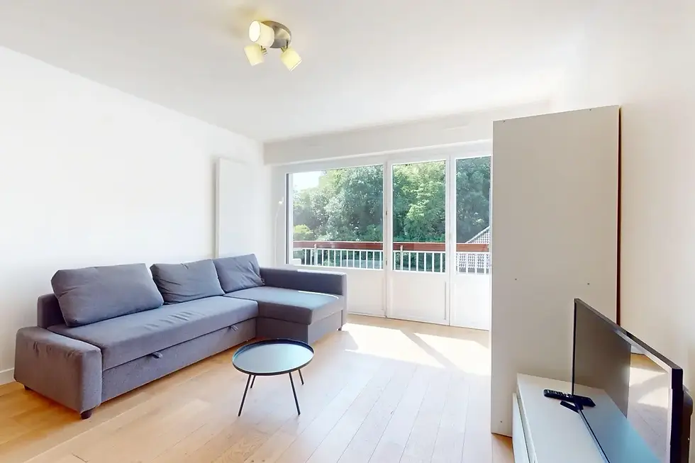 Salon lumineux de l’appartement Noma à Saint-Julien-en-Genevois, avec canapé d’angle gris et accès balcon verdoyant.