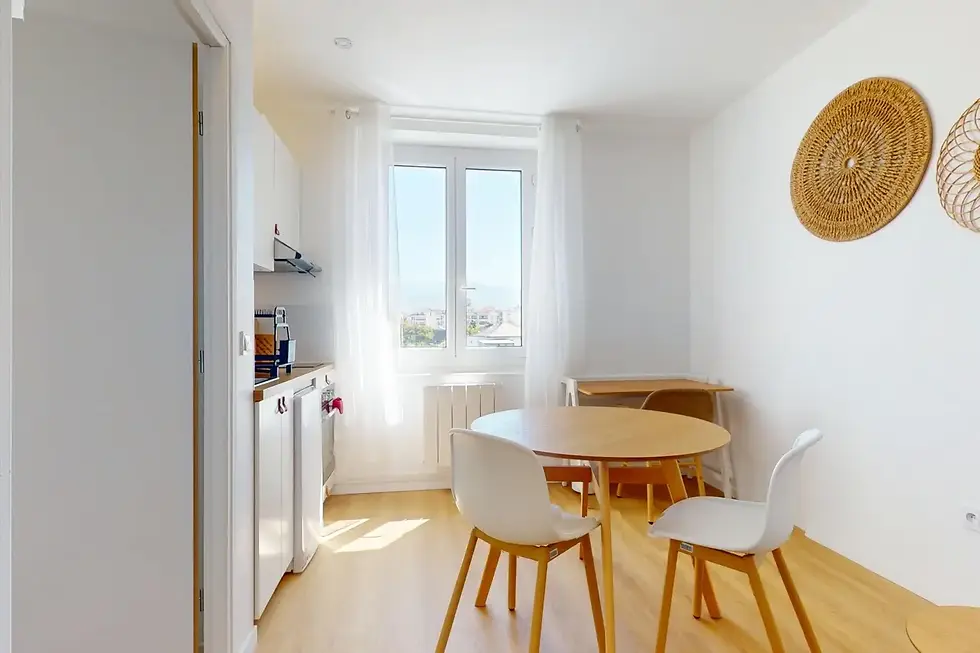 Appartement Orée à louer à Ambilly avec coin repas lumineux, table ronde en bois, kitchenette blanche et déco naturelle.