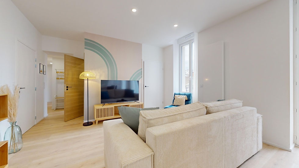 Salon lumineux avec canapé beige clair, coussins bleus et gris, meuble TV en bois clair, télévision à écran plat, fauteuil bleu avec coussin jaune, lampadaire en rotin. Colocation Colisée à Annemasse