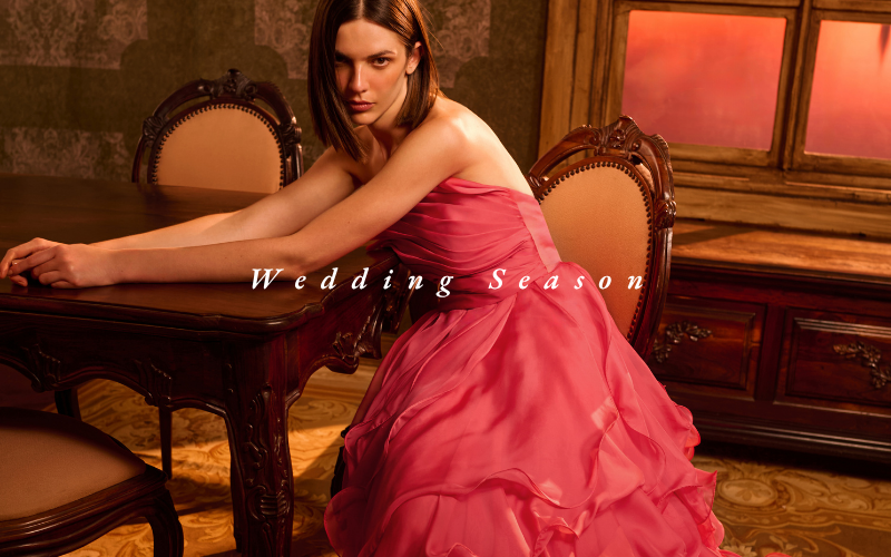 Mulher em vestido rosa sentada à mesa de madeira, fundo com janela laranja ao pôr do sol e texto "Wedding Season", atmosfera elegante.