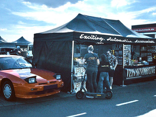 Impression von einer Automobil-Veranstaltung: Ein Infostand für Fahrzeugzubehör neben einem klassischen roten Sportwagen unter freiem Himmel.
