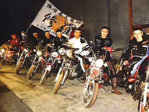 A line of Bōsōzoku members on modified motorcycles at night, with a large flag featuring Japanese characters in the background.