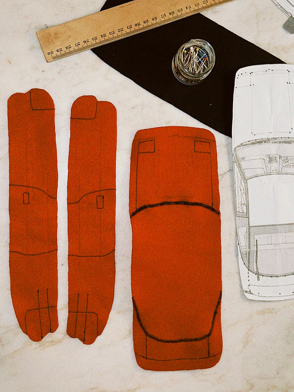 View of the fully prepared felt parts for a DIY fabric car: red side panels and roof section with precise black decorative stitching, as well as a paper template to check the proportions.
