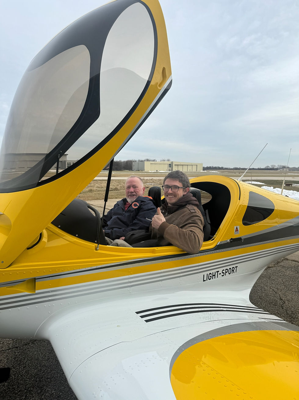 Two smiling students seated inside a bright yellow Bristell light-sport aircraft with the canopy open at the airport ramp, re