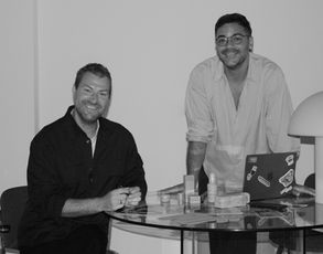 A black and white photo of Egon and Gerard, co-founders of RuMU Beauty, standing at a table with their products.