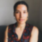 A photo of Ora Ozer, a person with tanned skin and dark textured hair that is tied up. They wear a top with a red floral pattern and hoop earrings.