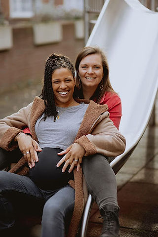 A photo of Tash and Marthe Koster-Thomas, a lesbian couple, sat at the foot of a slide. Tash has brown skin, long dark locs and is wearing a jacket, t-shirt and pregnancy yoga pants. is sat in front of Marthe, with her hands on her pregnant stomach. Marthe has pale skin and shoulder length brown hair. Both are smiling at the camera.