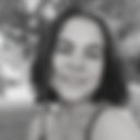 Foto em preto e branco de close do rosto de uma mulher parda, cabelos castanhos escuros lisos divididos ao meio, até os ombros, olhos pequenos, nariz regular, lábios finos em sorriso que mostra os dentes. Rosto de frente para a câmera e ombros de perfil à esquerda da tela. Fundo em cinza. Fim da descrição