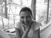 Foto em preto e branco de uma mulher negra, cabelos crespos curtíssimos, camiseta branca, apoiando a cabeça com a mão direita sob o queixo. Fundo em plano aberto onde se vê a rede onde ela está sentada, uma mesa com alguns livros e um quintal com plantas. Fim da descrição.