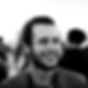 Foto em preto e branco de close do rosto de um homem branco, cabelos, barba e bigode curtos e pretos. Ele está sorridente. Fundo em plano aberto onde se vê algumas pessoas em ambiente branco. Fim da descrição