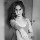 "Foto em preto e branco de uma mulher branca de cabelos longos castanhos e lisos. Ela está sentada e apoia o braço direito esticado sobre o joelho direito. Usa camiseta regata de alças fininhas. Está escorada numa porta escura.
Fim da descrição"