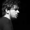 Foto em preto e branco, close de rosto de uma homem branco de perfil lateral direito, olhando para a direita da tela. Cabelos castanho escuros não muito curtos, barba e bigode rasos, camiseta preta. Fundo preto. Fim da descrição