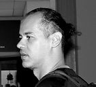 Foto em preto e branco em close do perfil esquerdo de um homem negro de cabelo curto amarrado em coque atrás da cabeça com lateral raspada. Veste camiseta preta. Ao fundo, parede branca com uma tela de tv preta desligada. Fim da descrição