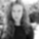 Foto em preto e branco de uma mulher branca, cabelos cacheados, divididos à esquerda da tela, soltos até abaixo dos ombros. Usa camiseta preta. Ao fundo, plantas. Fim da descrição