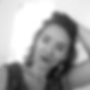 Foto em preto e banco em close de uma mulher branca, cabeça inclinada para a direita da imagem, braço esquerdo segurando o cabelo para trás da cabeça. Usa grandes brincos aparentes nas orelhas. Veste camiseta regata. Fundo em branco. Fim da descrição