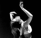 Foto em preto e branco em close de busto de uma mulher branca com o corpo direcionado para a direita quase de costas para a tela.
Cabelos castanho claros longos lisos, amarrados em rabo de cavalo, blusa folgada branca. A cabeça está jogada para trás, olhos fechados e rosto virado para cima, braços arqueados para cima, mãos soltas como quem dança. Fundo totalmente em preto. Fim da descrição.