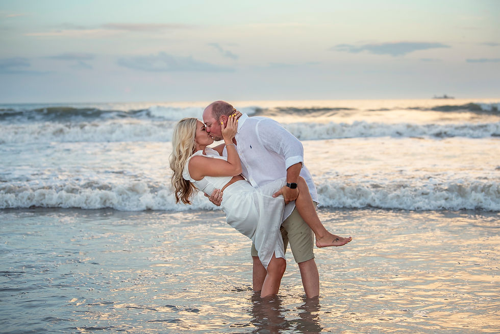 A couple kisses passionately in the ocean at sunset. The man lifts the woman, both in white attire, with gentle waves in the background.