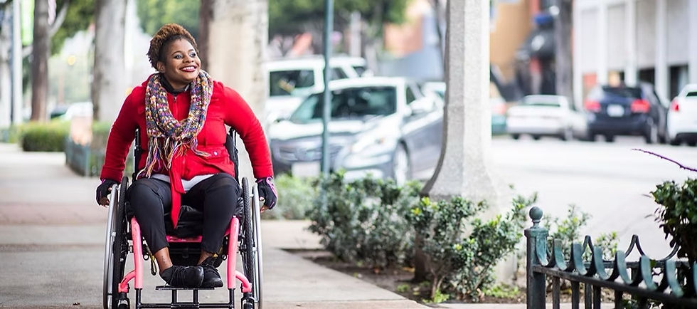 Woman in a wheelchair wearing a red coat and colorful scarf, smiling while moving down a city sidewalk. Cars and trees line the background.