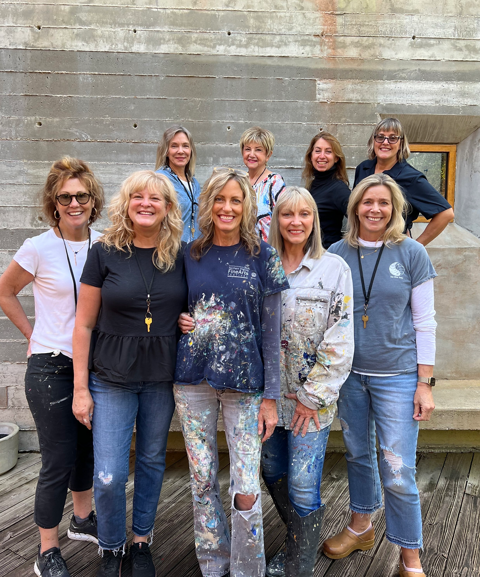 A group of nine smiling women posing outdoors, wearing casual and paint-splattered clothes against a concrete wall, exuding joy and creativity.