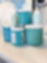 Jars of blue and teal paint stacked on a table, with a textured abstract painting in the background. Calm, creative studio vibe.