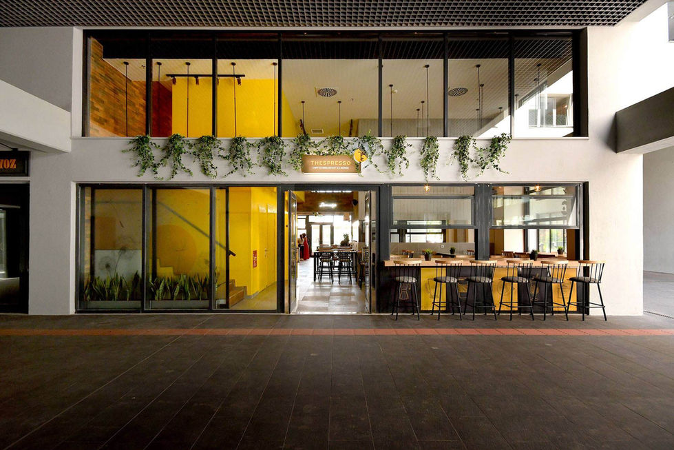 Exterior view of Thespresso café with pendant lights visible inside, modern lighting design complementing the yellow walls and greenery façade.