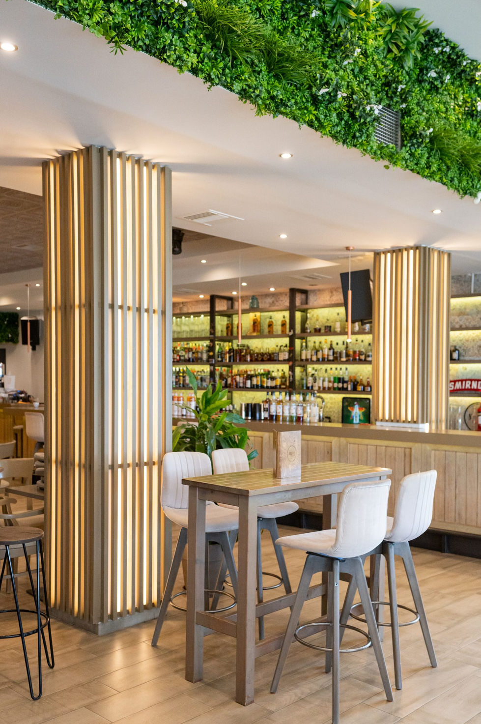 Close-up of illuminated vertical wooden slat columns next to a high table and bar stools, adding architectural rhythm to the bar interior.