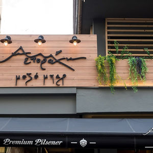 Exterior view of the meze bar with wooden signage, illuminated wall lamps, and cascading greenery from a balcony planter.