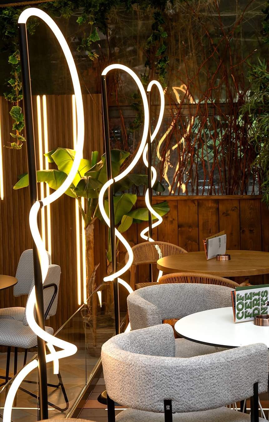 Curved sculptural LED light installations in front of a wooden wall with vertical linear lighting and lush plants.