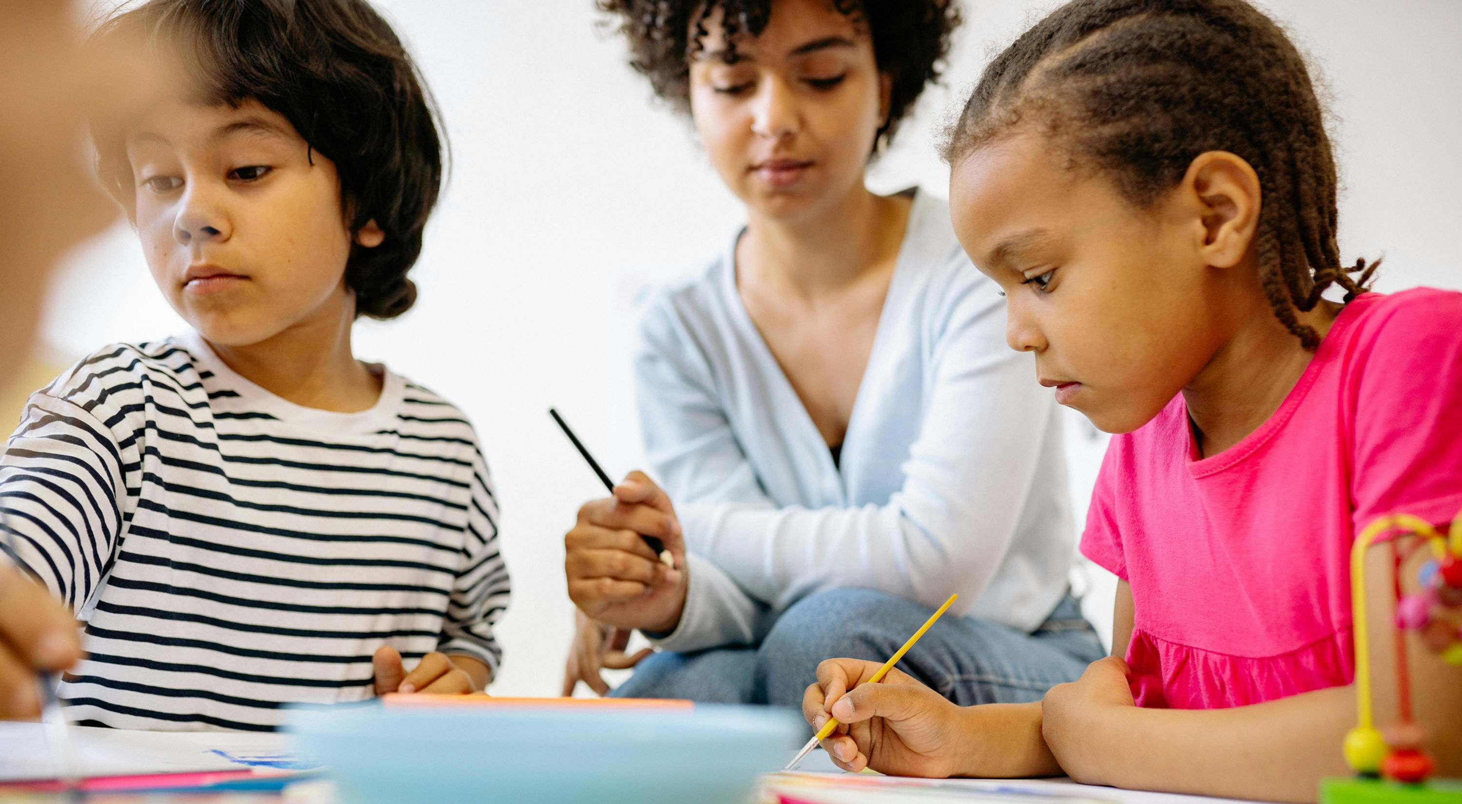 Un enfant au premier plan qui tient un pinceau et un adulte assis au second plan, crayon en main, qui regarde ce que fait l'enfant