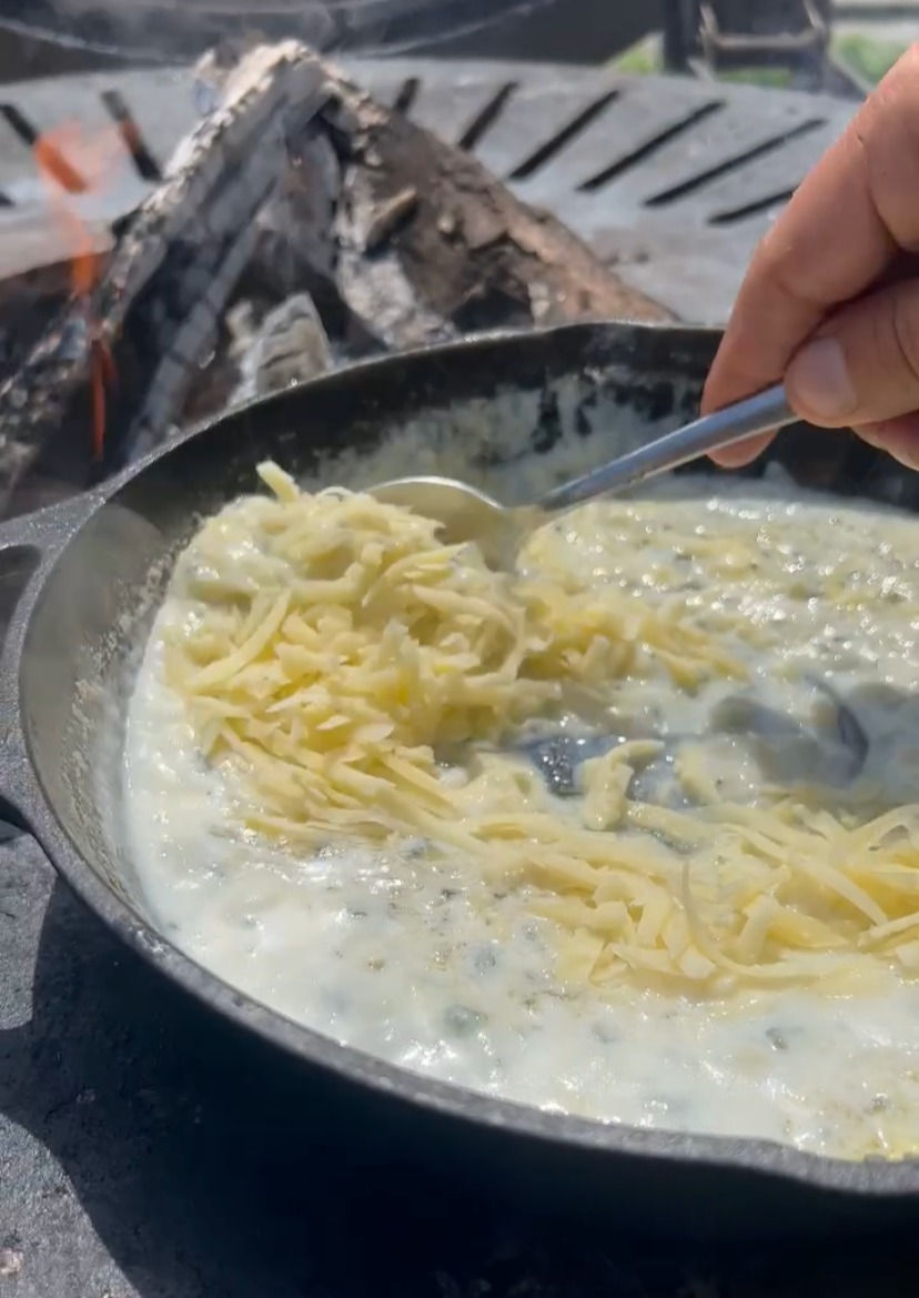 grated gruyere cheese being stirred into a roux in a cast iron pan on the plancha