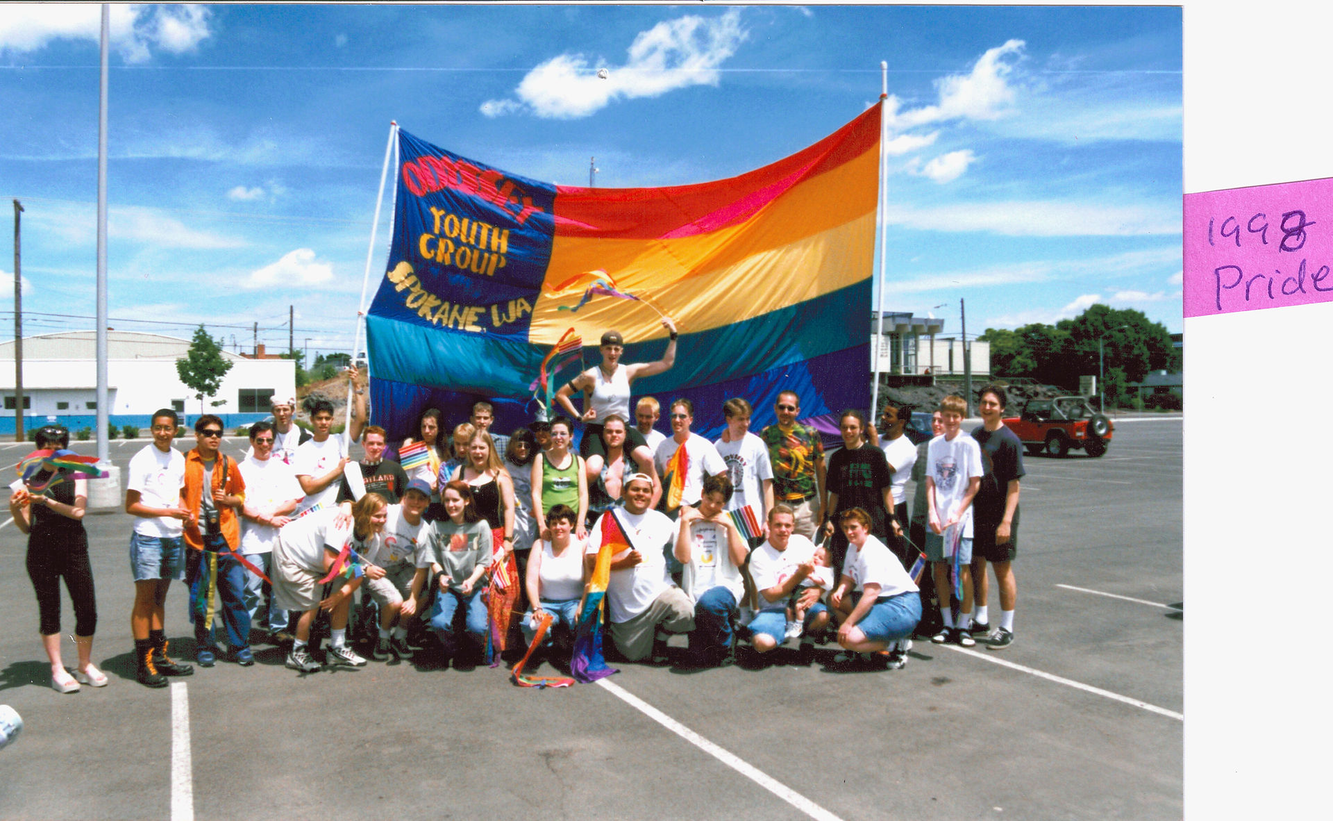 OYM at Pride (c. 1998). A large group of Odyssey youth pose in front of a large pride flag with "Odyssey Youth Group / Spokane, WA" written on it. Other smaller flags are held by a few of the members. Based on the amount of jean shorts present and the blue sky, it looks like a nice summer day.