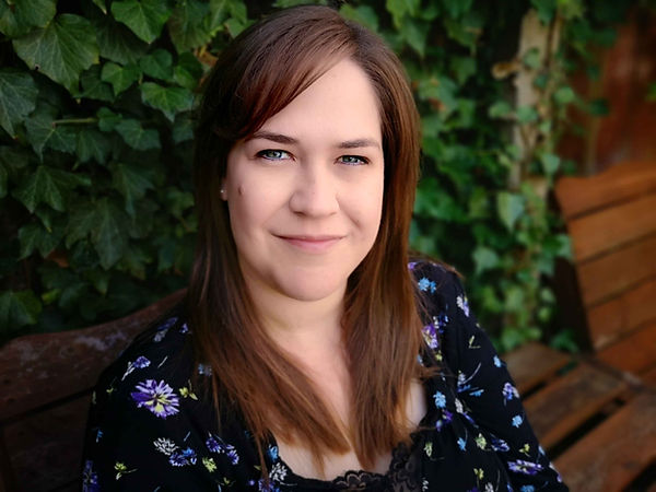 Katie Manasse - a woman in her mid thirties with long brown hair, sitting on a bench with a leafy background, smiling
