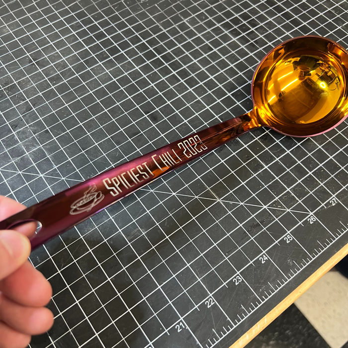 A shiny, red ladle with a gold bowl lies on a black cutting mat, the handle engraved with "Spiciest Chili"