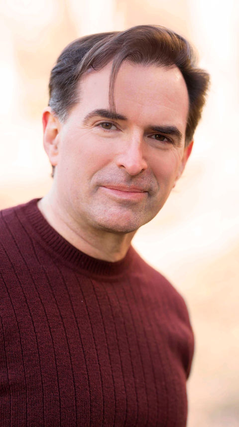 Commercial headshot of John Stanisci smiling with the sun behind him in a burgundy crew neck sweater