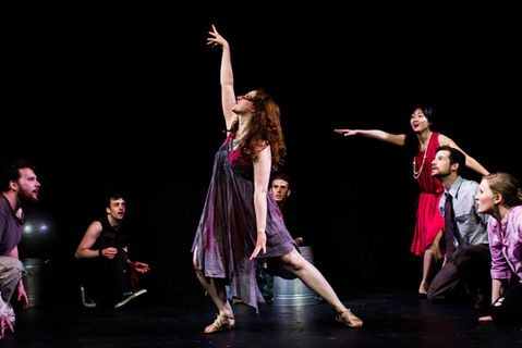 Ashley Ford in a colorful tattered dress stretches her arms to the sky the ensemble kneed around her looking up