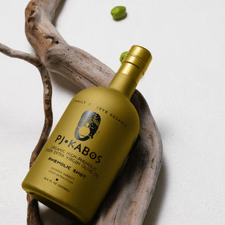 PJ Kabos Family Reserve Organic Phenolic Shot high-phenolic extra virgin olive oil gold bottle resting on driftwood with green olives on a white background.