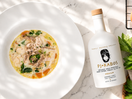 A bowl of Avgolemono Chicken Soup with a bottle of PJ Kabos High-Phenolic Olive Oil sitting ready to eat on a table.
