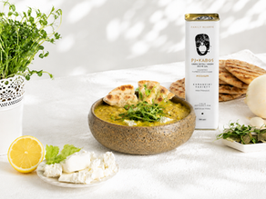 Bowl of Mediterranean yellow split pea soup garnished with feta cheese, fresh greens, and extra virgin olive oil, served with pita bread, lemon, onion, and a tin of PJ Kabos Family Reserve Medium olive oil on a bright white background.