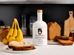 Freshly baked banana bread sitting on a table surrounded by its primary ingredients: overripe bananas and olive oil.