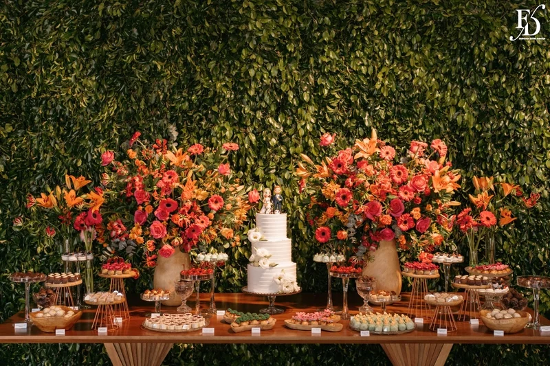Mesa de doces de casamento com decoração de flores coloridas laranja e rosa e muro inglês